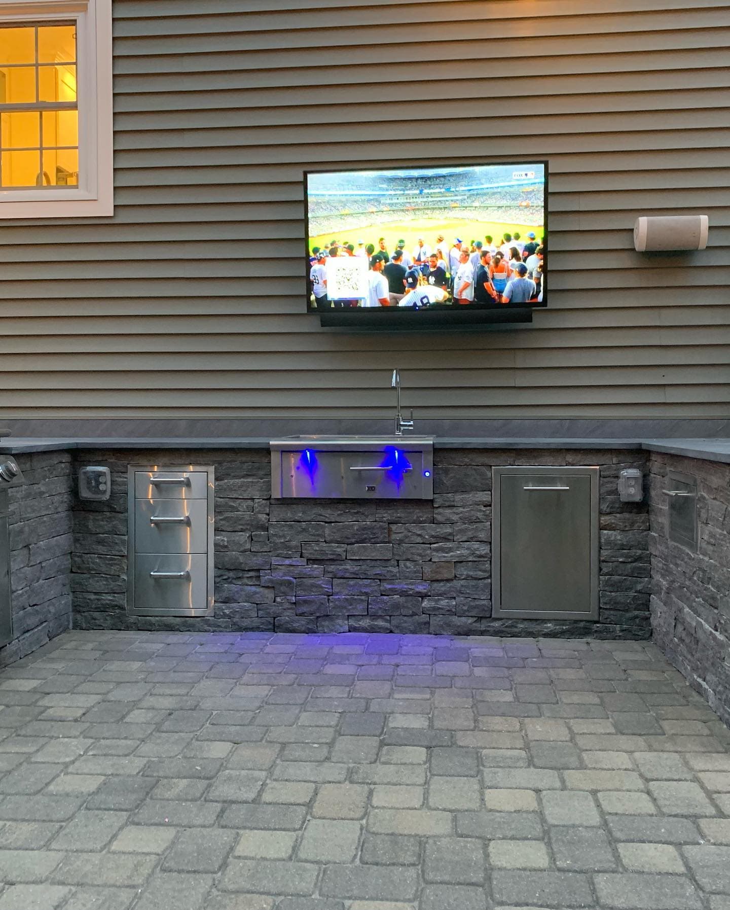 Stacked-stone outdoor kitchen with lit drawers and a TV mounted above